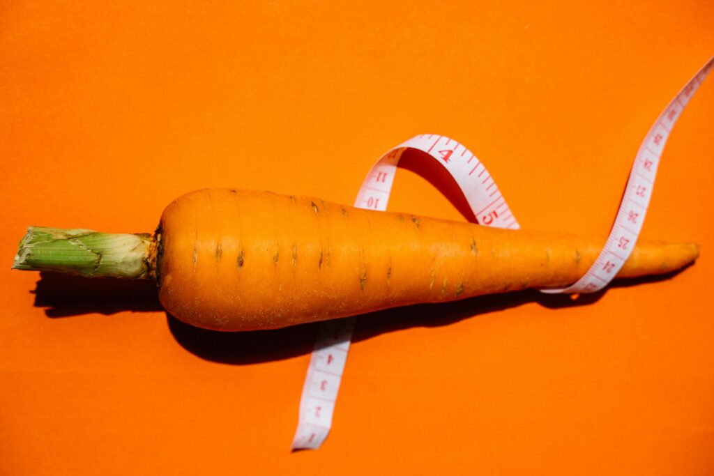 A vibrant image of a fresh carrot wrapped in a measuring tape on a bright orange background symbolizing health and nutrition.