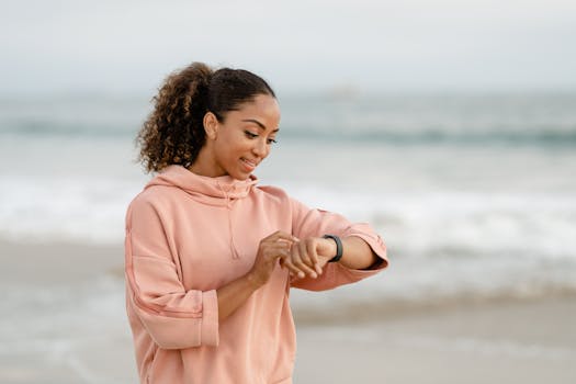 Smiling woman in a pink hoodie checks her smartwatch by the ocean.
