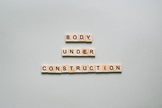 Wooden scrabble tiles spell 'Body Under Construction' on a minimalist white background, conveying motivation.