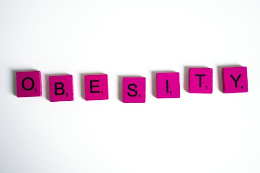 Purple Scrabble tiles spelling 'obesity' on a white surface, conceptual still life.