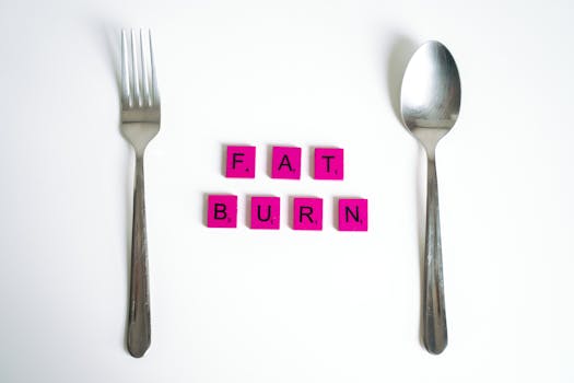 Conceptual art featuring the words 'Fat Burn' using Scrabble tiles between a fork and spoon on a white background.