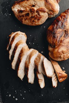 Sliced and whole grilled chicken breasts on a dark slate surface, seasoned perfectly.