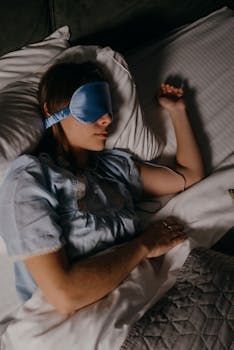 Woman peacefully sleeping with a blue eye mask in a cozy bed setting, promoting restful sleep.