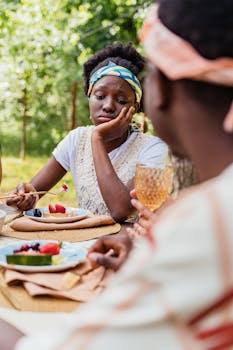 Woman looking contemplative outdoors, holding a fork with fruit at a picnic table.