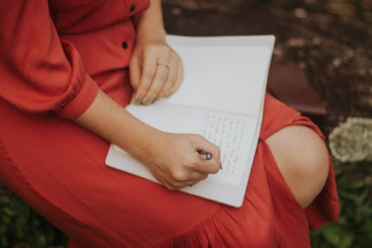 A woman in a red dress writing in a journal, outdoors over a leafy background.