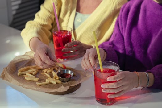 Two individuals sharing french fries with ketchup and red drinks indoors.