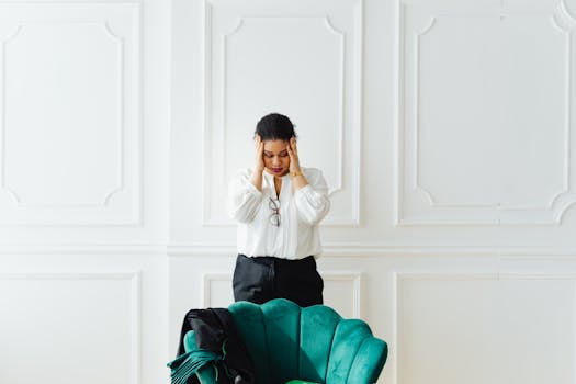 A woman holds her head in stress, standing indoors next to a green chair.