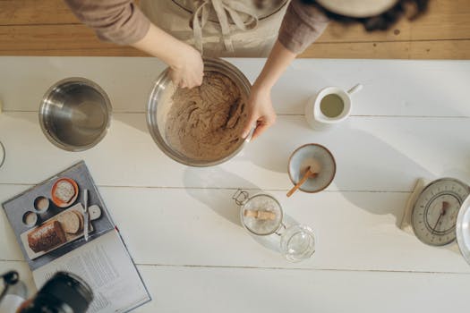 A close-up of hands mixing batter in a bowl among baking ingredients on a wooden table.