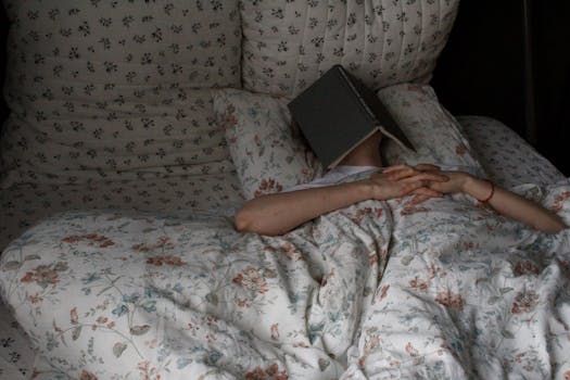 Woman relaxing in bed with book on face, floral bedding, creating a cozy vibe.