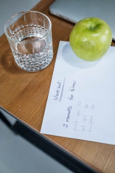A refreshing glass of water with a green apple and workout plan on a wooden table.