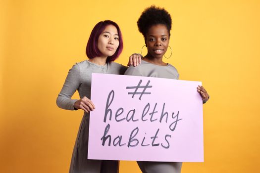 Two women holding a sign with '#healthy habits' on a vibrant yellow background.