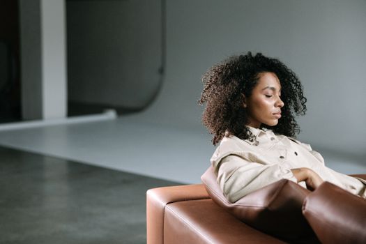A serene black woman with curly hair rests on a brown sofa in a minimalist indoor setting.