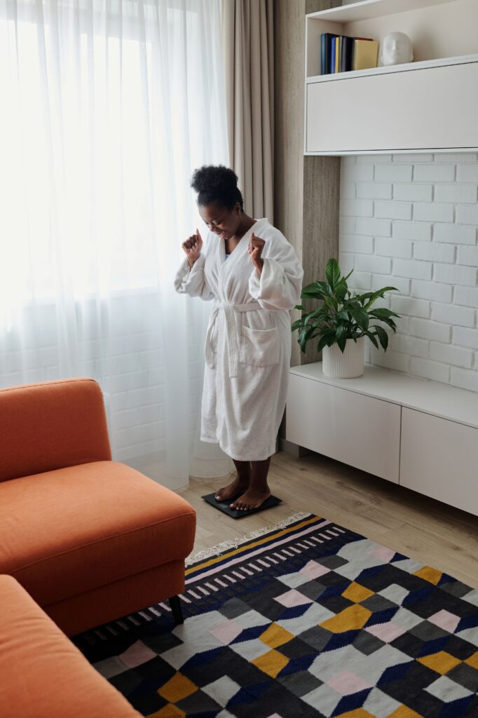 African American woman in a bathrobe, happily checking weight in a modern home interior.