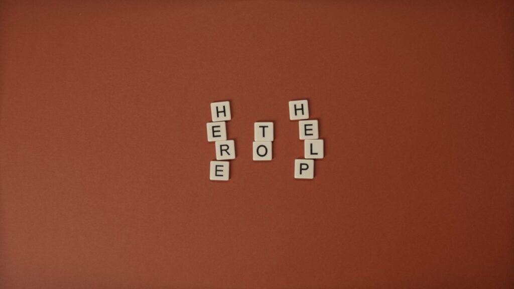 Flat lay of Scrabble tiles spelling 'Here to Help' on a brown background with copy space.
