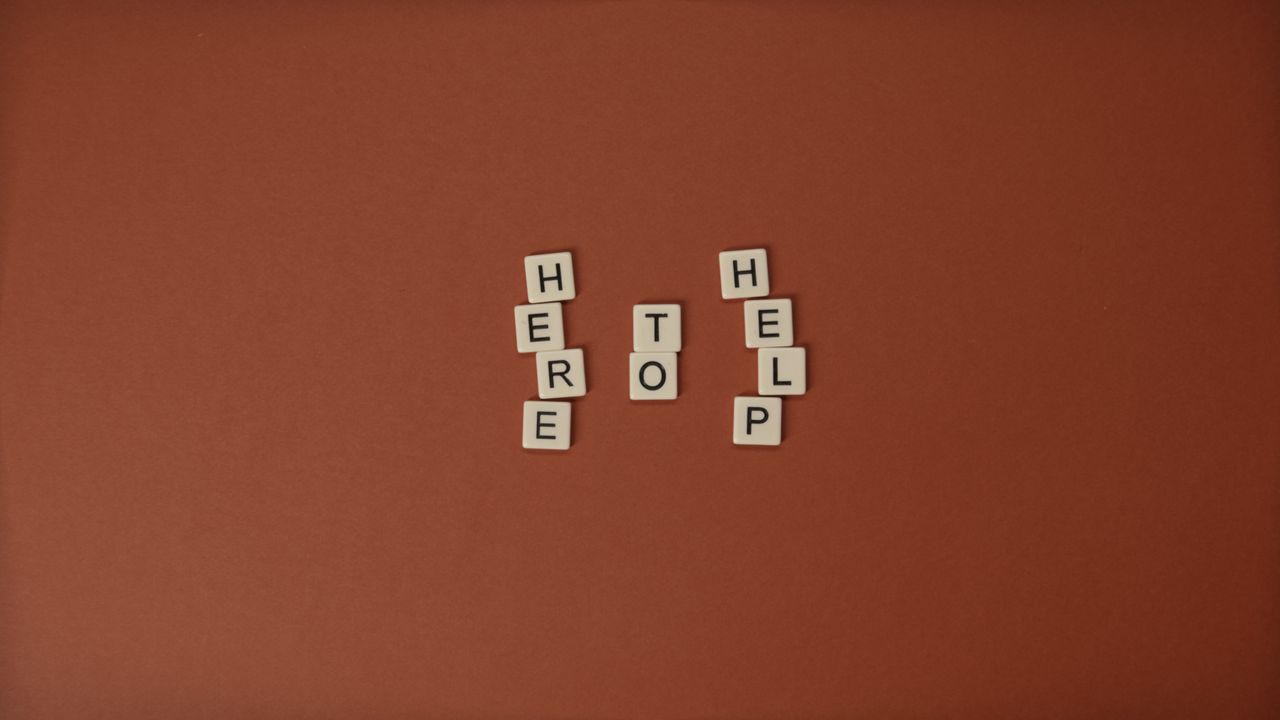 Flat lay of Scrabble tiles spelling 'Here to Help' on a brown background with copy space.